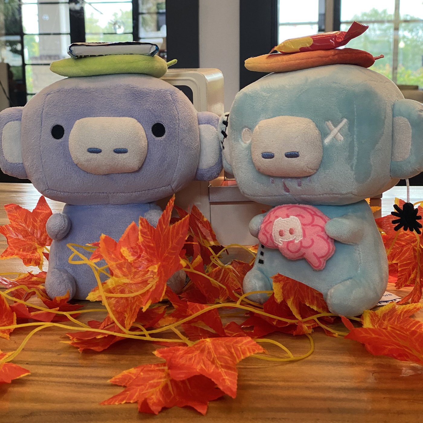 Two Wumpus plushies, one normal and one zombie with a brain, on a table of autumn leaves